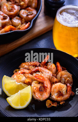 Skillet roasted jumbo shrimp with sliced garlic and spices on a black plate. Closeup. A glass of beer and a skillet in the background.