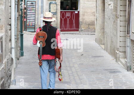 Musician wearing a straw fedora hat, carrying a mandolin and ukelele, walking down a cobbled street towards a small cafe Stock Photo