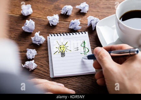 Person Drawing Light Bulb And The Financial Gain On Notepad Near Crumpled Paper On Wooden Desk Stock Photo