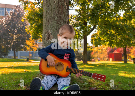 Small boy holding acoustic guitar while sitting leaned on a tree Stock Photo