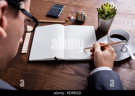High Angle View Of Businessman Hand With Pencil On Notebook At Wooden Desk In Office Stock Photo
