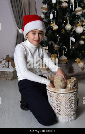 Close up of smiling mischievous boy in white sweater and Santa red hat. Boy is sitting near decorated Christmas tree and taking out the brown teddy be Stock Photo