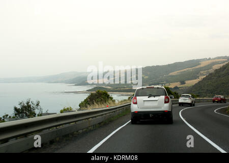 Cars driving along a winding coastal road on an overcast day with the ocean to one side and mountains on the other Stock Photo