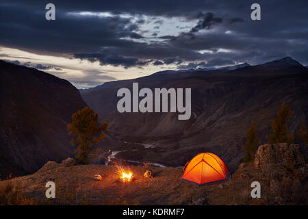 A illuminated tent and campfire in mountains in dawn Stock Photo