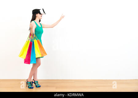 pretty attractive lady holding many shopping bags standing on wood floor and wearing 3d technology vr device using hands catching virtual image in whi Stock Photo