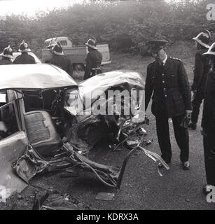 1960s, Buckinghamshire, UK, poiice and firemen with helmets at the scene of a rural road accident and a severely smashed up motorcar lying on the road.