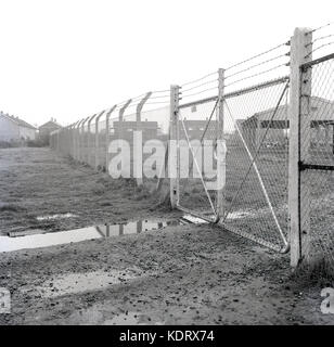 1960s, historical, England, UK, chain link fencing with barbed wire on top and locked gates protect and block any illegal entry to an old industrial storage yard on the edge of a large rural housing estate. Stock Photo