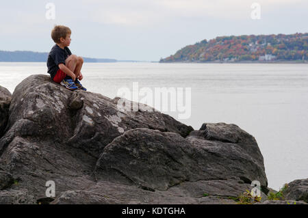 A little boy (5 yrs old) sitting alone on rocks by the River St Lawrence at Cap Rouge near Quebec City, looking thoughtful