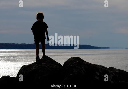 A little boy(5 yrs old) silhouetted standing on rocks on the St Lawrence river shore at Cap Rouge near Quebec City, Canada Stock Photo