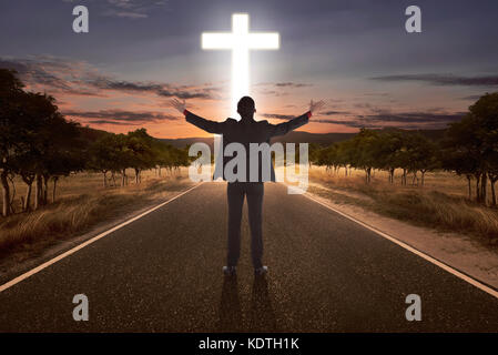 Portrait of man raising hand while praying to god with bright cross at the end of the road Stock Photo