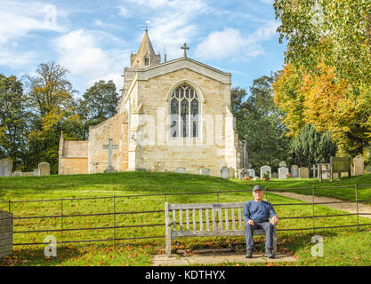 Retired, smiling man relaxes on a wooden bench in front of the east end of the medieval christian church of St Andrew, dating from the thirteenth cent Stock Photo