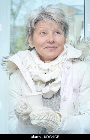 Gray haired senior woman in winter fur jacket and knitted mittens enjoing a drink. Soft filter. Blue toning effect. Stock Photo