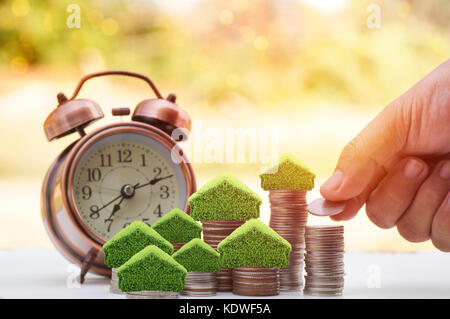 hand put money on stacked of coins to save for house, small tree and home on pile with alarm clock on wood table with sunlight background, concept as  Stock Photo
