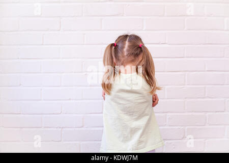 Female child with back to viewer and arms folded in grumpy or sad pose against white brick wall. Emotional or unhappy kid concept Stock Photo