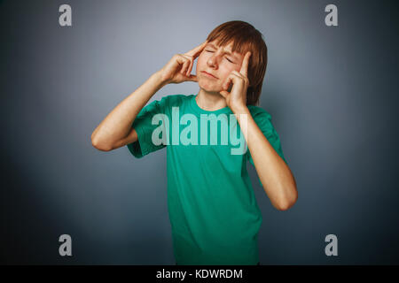 boy teenager European appearance brown hair in a turquoise t-shirt pressed his fingers to his temple on a gray background, thinking, thought cross pro Stock Photo
