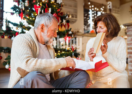 Senior couple sitting on the floor in front of illuminated Christmas tree inside their house giving presents to each other, woman unwrapping her gift. Stock Photo