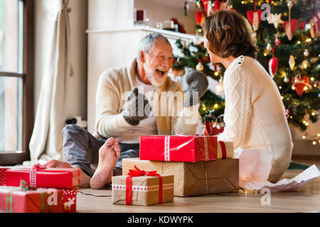 Senior man sitting on the floor in front of illuminated Christmas tree with his wife. The man is trying on knitted mittens. Stock Photo