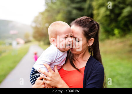 Crying little boy with his mother in nature. Young woman holding her toddler boy. Summer time. Stock Photo