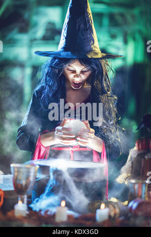 Portrait of young angry witch with awfully face in creepy surrounding watching the future in magic ball above boiling potion. Stock Photo