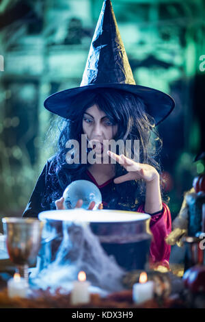 Portrait of young witch with awfully face in creepy surrounding watching the future in magic ball by the boiling potion. Stock Photo