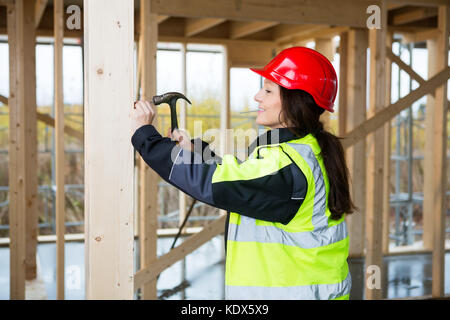Side view of female carpenter in reflective jacket hammering nail in wood at construction site Stock Photo