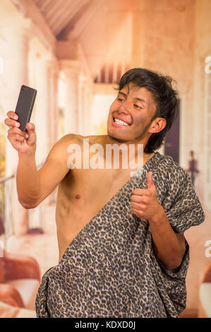 Close up of funny prehistoric man doing funny faces to the camera, taking a selfie with his cellphone and thumbs up, in a blurred background Stock Photo