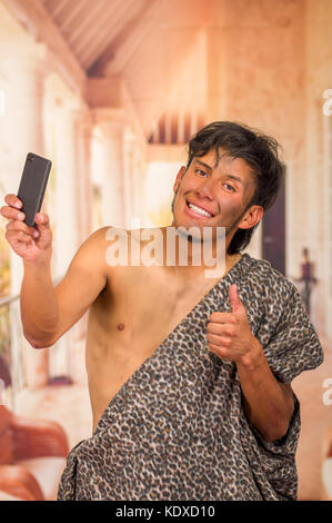 Close up of funny prehistoric man doing funny faces to the camera, taking a selfie with his cellphone and thumbs up, in a blurred background Stock Photo