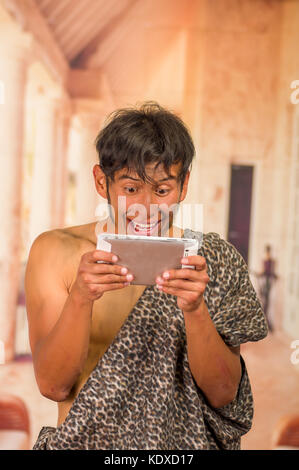 Close up of funny prehistoric man doing funny faces to the camera, holding in his hands a tablet, in a blurred background Stock Photo