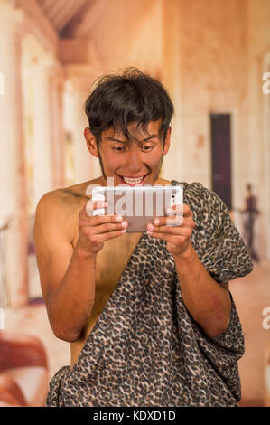 Close up of funny prehistoric man doing funny faces to the camera, holding in his hands a tablet, in a blurred background Stock Photo