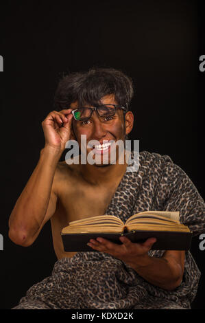 Close up of funny prehistoric man smiling to the camera, wearing glasses and reading a book in a black background Stock Photo