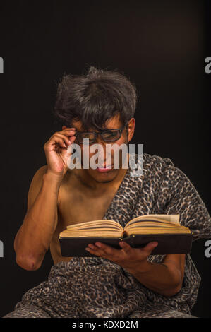 Close up of funny prehistoric man smiling to the camera, wearing glasses and reading a book in a black background Stock Photo