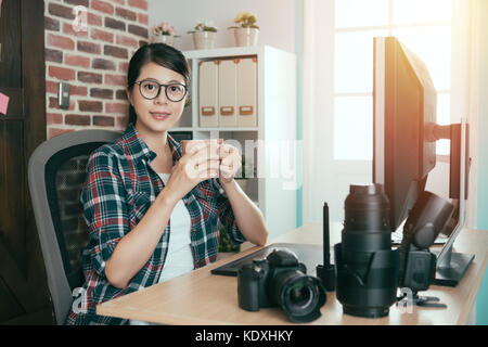 happy attractive woman photographer face to camera smiling and sitting on office desk working drinking hot espresso coffee resting relaxing at afterno Stock Photo