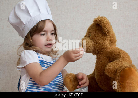 Portrait Adorable little girl in chef hat. baby feeds a toy bear Stock Photo
