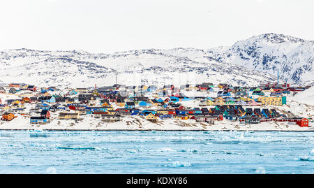 Arctic city panorama with colorful Inuit houses on the rocky hills covered in snow with snow and mountain in the background and blue  icebergs in a fo Stock Photo