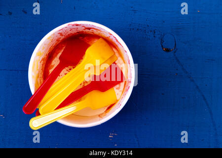 Empty messy ice cream paper cup with yellow and red plastic spoons isolated on blue painted wood from above. Stock Photo