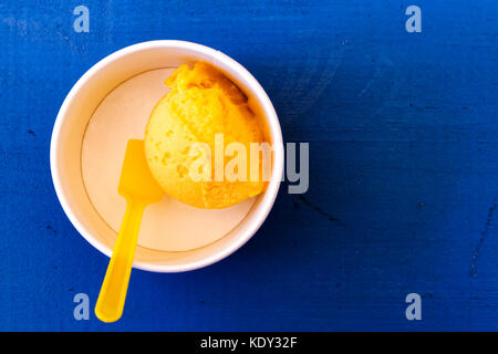 Scoop of mango ice cream in paper cup with plastic spoon isolated on blue painted wood from above. Stock Photo