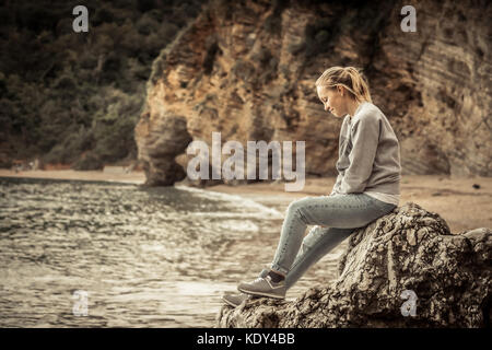 Pensive lonely young woman traveler relaxing on a big cliff stone on the beach looking at wild mountain scenery in retro vintage style Stock Photo