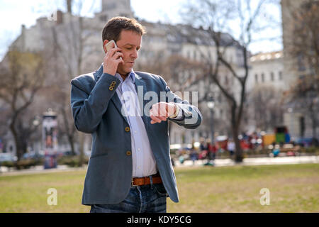 A middle age businessman standing in a park while talking on his phone and checking the time on his watch Stock Photo