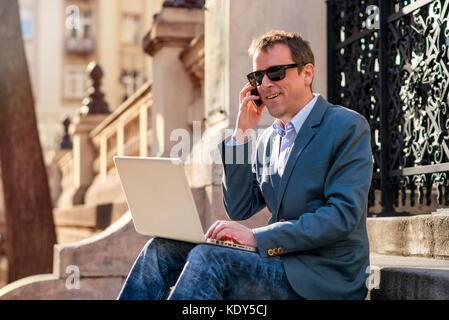 A middle age businessman siting on the stairs with a laptop while talking on the phone and wearing sunglasses Stock Photo