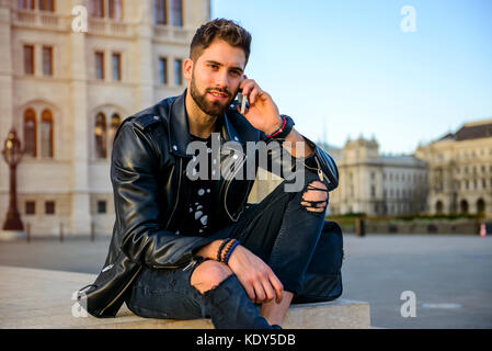 A handsome young hipster man sitting on the stairs on a square while talking on his smartphone Stock Photo