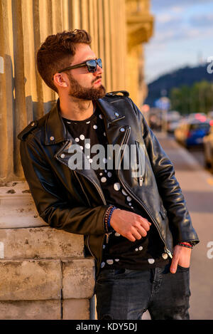 A handsome young hipster man leaning to the pillar of a house on a sunny street and relaxing with his sunglasses on Stock Photo