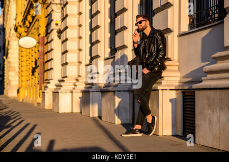 A handsome young hipster man leaning to the wall of a house on a sunny street while talking on his smartphone Stock Photo
