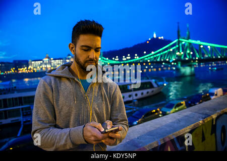 A handsome young sportsman using his phone while standing in the city lights on the riverside at night Stock Photo