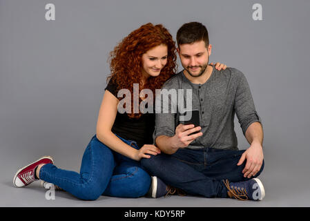 A beautiful young couple sitting on the ground and looking at a smartphone and smiling Stock Photo