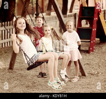 Laughing girls and boys in school age swinging on children's playground outdoors Stock Photo