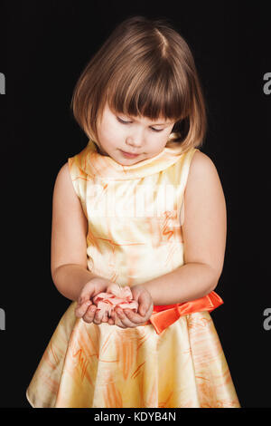 Little blond girl in yellow dress holds pink rose flower made of feathers on black background, vertical studio portrait Stock Photo