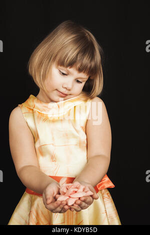 Little Caucasian girl in yellow dress with pink rose flower made of feathers on black background, vertical studio portrait Stock Photo