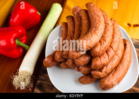 Stack of deliciously smoked handmade Swedish Isterband sausages with natural casing. Here on white plate sided by red bell pepper and leek. Stock Photo