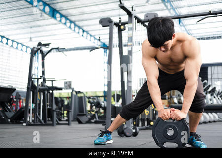 young man bodybuilder execute exercise in fitness center. male athlete lift heavy weight barbell plate in gym. sporty asian guy working out in health  Stock Photo