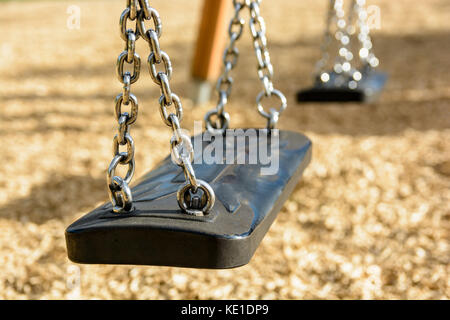 Close-up view of a still child's swing in black plastic in a wood chips covered playground with chrome chains and drop shadow. Stock Photo